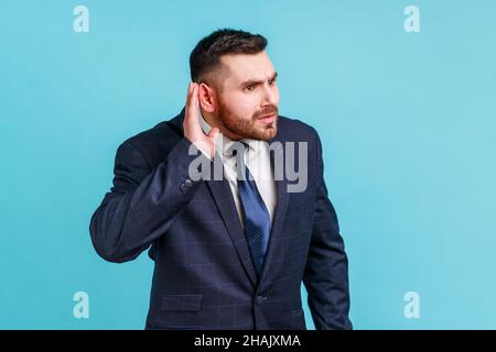 Je ne peux pas vous entendre.Portrait de l'homme de brunette nosé avec la barbe en costume essayant d'entendre des ragots, tenant la main près de l'oreille et écoutant attentivement.Studio d'intérieur isolé sur fond bleu. Banque D'Images