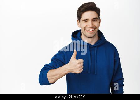 L'image d'un jeune homme souriant montre le pouce et se met à l'image de l'appareil photo, heureux par smth, approuver ou aimer, compliment, fond blanc Banque D'Images