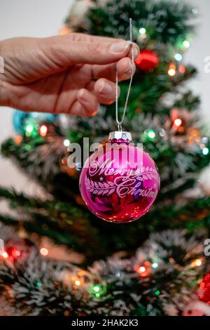 Main de femme décorant un arbre de Noël avec un ornement de fête rose vintage avec un signe de Joyeux Noël.Des lumières de fête floues en arrière-plan. Banque D'Images