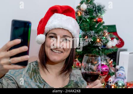 Une femme porte un chapeau de Père Noël, devant l'arbre de Noël, tenant un verre de vin et parlant à sa famille via un appel vidéo sur son smartphone. Banque D'Images
