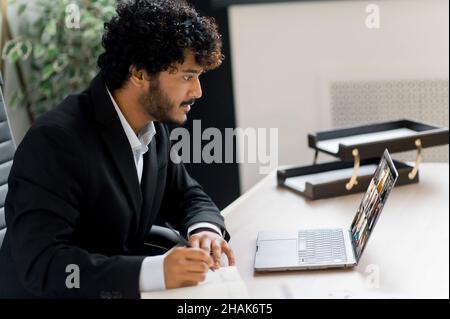 Jeune adulte ciblé homme d'affaires indien réussi en costume formel assis à la table dans le bureau, parlant sur la conférence en ligne via ordinateur portable avec des collègues, prenant des notes, regardant l'écran Banque D'Images