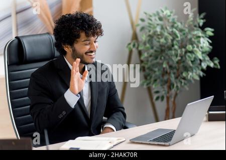 Sympathique, jeune adulte heureux, intelligent, grand responsable indien, PDG ou homme d'affaires avec une barbe, assis à la table, accueille les employés d'un geste de la main lors d'une conférence en ligne, souriant Banque D'Images