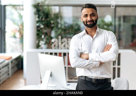 Portrait d'un beau chef d'entreprise indien, PDG, directeur général, spécialiste INFORMATIQUE, séduisant, confiant, intelligent et influent, dans une chemise blanche, debout près de la table au bureau, regarde le sourire de l'appareil photo Banque D'Images