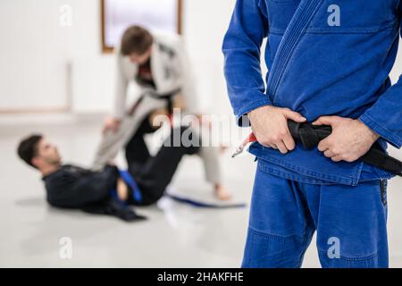 Gros plan sur la section médiane de l'athlète masculin inconnu du caucase bjj brésilien jiu-jitsu ceinture noire debout sur le tapis à l'académie tenant sa ceinture pendant le traa Banque D'Images