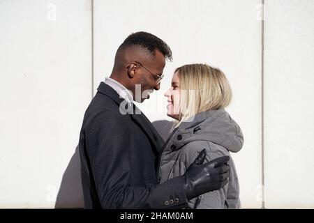 Homme noir en lunettes et manteau embrassant sa petite amie blonde en manteau par temps ensoleillé. Banque D'Images