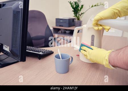 Nettoyage des appareils de cuisine dans le bureau, main d'homme dans des gants jaunes essuie la bouilloire avec une éponge Banque D'Images