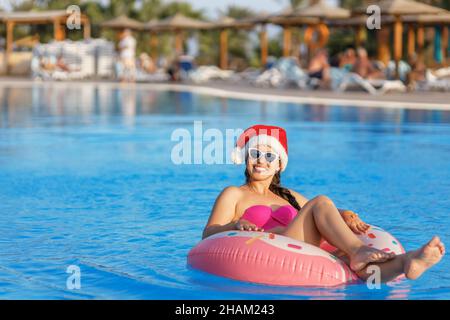 belle jeune femme en chapeau de père noël et lunettes de soleil se détendant sur l'anneau gonflable dans la piscine Banque D'Images