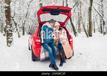 Couple amoureux assis dans le coffre de voiture ayant le thé chaud dans la forêt enneigée d'hiver.Homme qui verse la boisson du thermos.Les gens se détendent à l'extérieur lors d'un voyage en voiture Banque D'Images