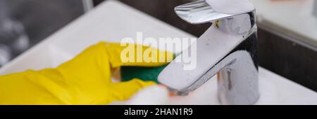 La personne portant des gants jaunes lave l'évier avec un chiffon et vaporise de la mousse nettoyante sur le robinet Banque D'Images