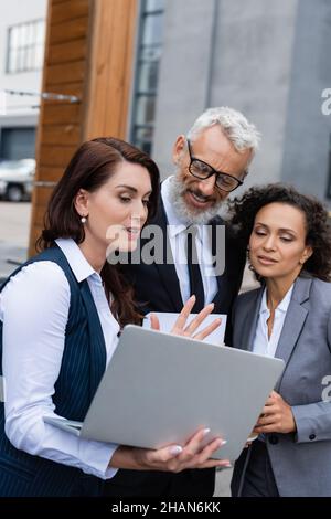 realtor montrant l'ordinateur portable à des partenaires commerciaux interraciaux positifs en plein air Banque D'Images