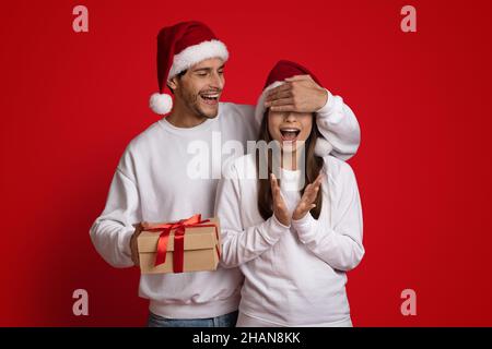 Joyeux jeune homme au chapeau de père Noël qui fait la surprise de Noël à une petite amie excitée Banque D'Images