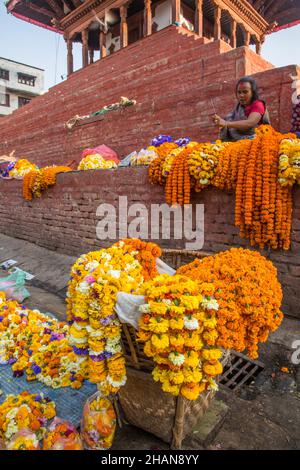 Une femme qui fait des guirlandes de fleurs pour des offrandes religieuses hindoues à vendre sur une plinthe de temple.Place Durbar à Katmandou, Népal. Banque D'Images