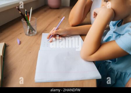 Gros plan rogné de fille primaire faisant des devoirs écrivant dans un livre avec un stylo avec une jeune mère assis à la table à la maison par fenêtre. Banque D'Images