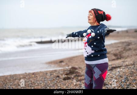 Femme âgée de 80s ans sur une plage s'exerçant en hiver.Elle porte un pull et un chapeau de Noël tout en faisant des sauts d'étoiles sur la côte sud, au Royaume-Uni Banque D'Images