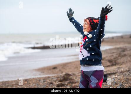 Femme âgée de 80s ans sur une plage s'exerçant en hiver.Elle porte un pull et un chapeau de Noël tout en faisant des sauts d'étoiles sur la côte sud, au Royaume-Uni Banque D'Images