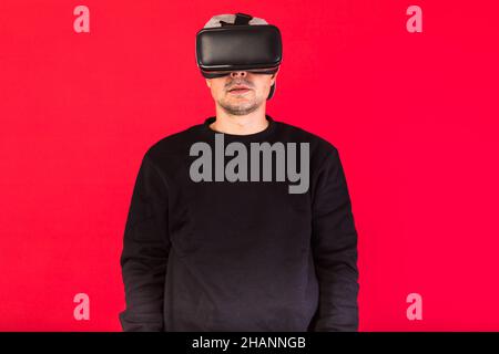 Jeune homme à barbe courte, portant un sweat-shirt noir, une casquette et des lunettes de réalité virtuelle stupéfait, regardant droit devant, sur fond rouge.Technologie, V Banque D'Images