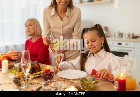 Femme millénaire mettant de la salade de légumes sur l'assiette de sa fille pendant le dîner de Noël avec la famille à la maison Banque D'Images