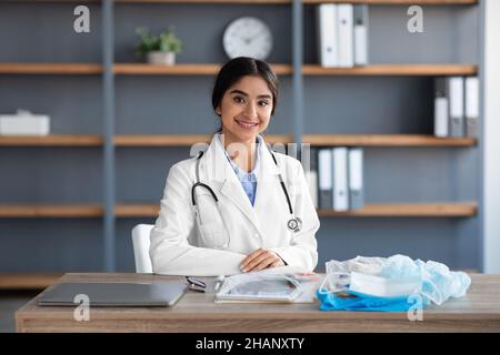 Souriante jolie jeune femme hindoue médecin en manteau blanc avec stéthoscope assis à la table avec beaucoup de masque de protection Banque D'Images