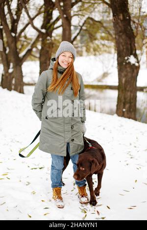 Jeune femme souriante avec chien brun labrador retriever en hiver chaud tenue marchant à l'extérieur dans un parc enneigé, plein corps regardant l'appareil photo Banque D'Images