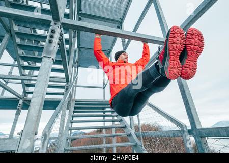 Un homme ajusté vêtu de vêtements sport rouge vif et de chaussures de course à pied accrochant la barre horizontale d'entraînement abs en levant les jambes.Entraînement physique sur l'envers en acier Banque D'Images