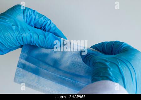 Main dans un gant en caoutchouc bleu tenant et contrôlant le fil du masque chirurgical à trois plis sur fond gris Banque D'Images