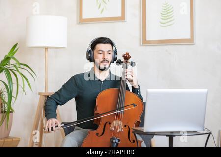 Jeune professeur confiant de musique dans des écouteurs jouant des instruments de musique à cordes pendant la leçon en ligne Banque D'Images
