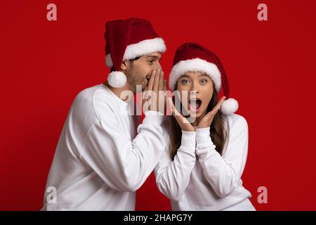 Jeune Guy à Santa Hat Whispering Christmas Secret à sa petite amie excitée Banque D'Images