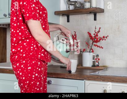 Femme en pyjama versant du thé à la maison table de cuisine le matin. Banque D'Images