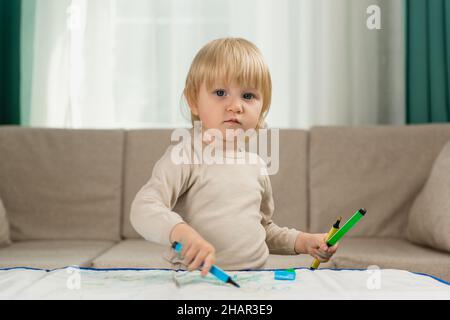 Une petite fille blonde en pyjama à la maison tire avec soin sur la table avec des marqueurs d'eau pour enfants Banque D'Images