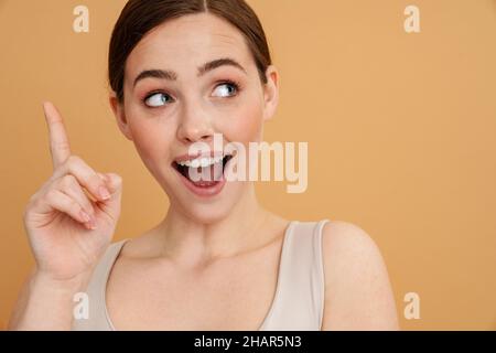 Gros plan d'une jeune femme souriante aux cheveux blancs et rouges, debout sur fond beige et pointant du doigt vers le haut Banque D'Images
