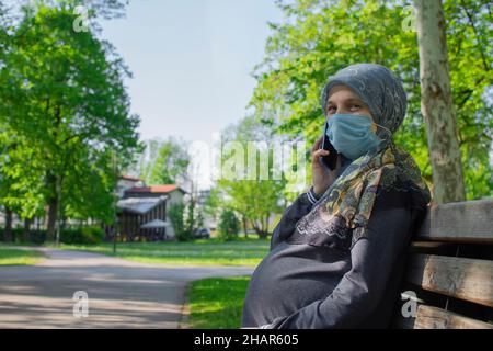 Profil de la femme enceinte musulmane portant un masque de visage parlant sur le smartphone Banque D'Images