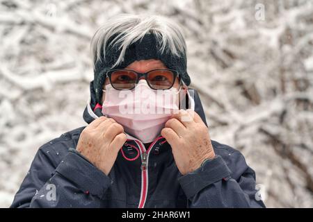 Femme âgée sénior avec cheveux gris portant un masque rose jetable de virus, hiver flou neige couverte arbres fond Banque D'Images