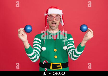 homme émerveillé en costume d'elf et chapeau de père noël. guy en lunettes de fête tient des boules de décoration de noël Banque D'Images