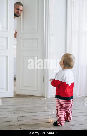 Jolie petite fille de bébé en costume de Santa debout dans la grande pièce lumineuse regardant Dad, heure de noël Banque D'Images