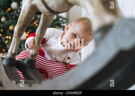 Petite fille en costume de Père Noël rouge regarde derrière un cheval en bois jouet dans la chambre avec arbre de Noël à la maison Banque D'Images