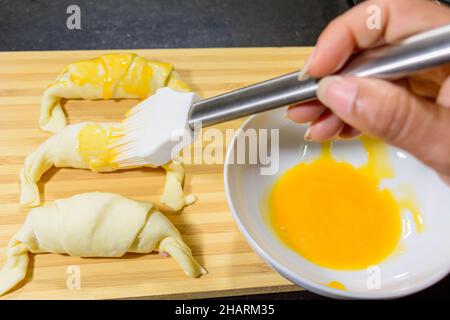 Chef peignant les croissants au jambon et au fromage avec du jaune d'œuf sur une planche en bois Banque D'Images