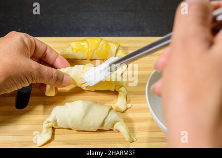 Photo d'un chef peignant les croissants au jambon et au fromage avec du jaune d'œuf sur une planche en bois Banque D'Images