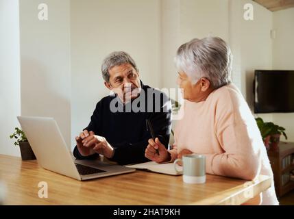 Couple de personnes âgées multiculturelles assis au comptoir de cuisine moderne avec ordinateur portable.En discutant du budget, la femme prend des notes dans un carnet avec un stylo. Banque D'Images