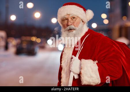 Portrait horizontal moyen de l'homme âgé portant le costume de Santa avec lunettes sac de transport avec cadeaux traversant la route le soir Banque D'Images