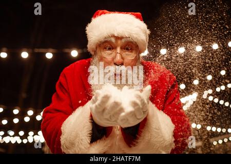 Portrait en gros plan d'un homme de grande garde espiègle portant le costume du Père Noël tenant la neige entre les mains et la soufflant à l'appareil photo Banque D'Images