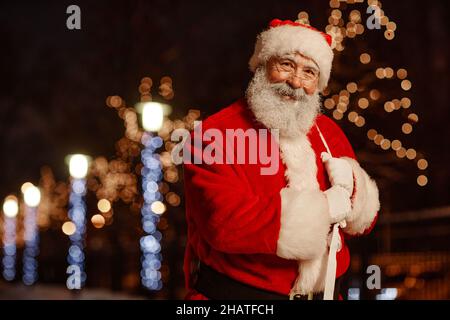 Portrait horizontal moyen du joyeux Père Noël debout à l'extérieur dans la rue décorée de guirlandes lumières souriant à l'appareil photo, espace de copie Banque D'Images
