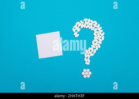 Point d'interrogation fabriqué à partir de pilules blanches et de comprimés avec petit papier mémo vierge, couché sur fond bleu. Industrie pharmaceutique mondiale et médicaments Banque D'Images