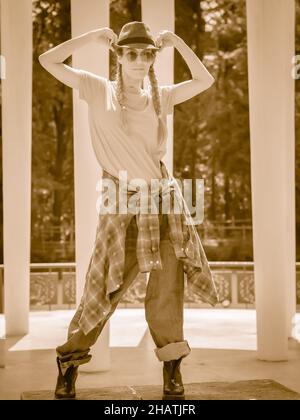 Une fille avec des queues de porc et un ensemble décontracté tient son chapeau au milieu des colonnes dans le belvédère du parc.Danseur de rue.Photographie monochrome Banque D'Images