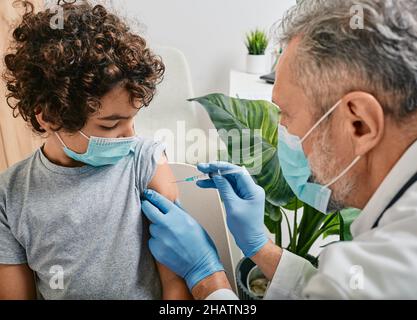 Vaccinations de routine pour un enfant.Un garçon aux cheveux bouclés portant un masque médical lorsqu'il est vacciné par un pédiatre mature à la clinique médicale Banque D'Images