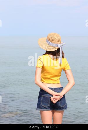 jeune fille skinny avec chapeau de paille et mains derrière son dos debout près de la mer en été Banque D'Images