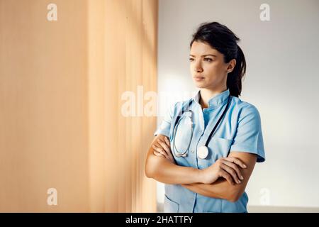 Personnel médical et service médical.Une jeune femme médecin debout avec les bras croisés dans son bureau et regardant par la fenêtre.Elle s'inquiète d'abo Banque D'Images