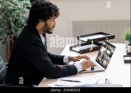 Jeune adulte sérieux concentré, chef d'entreprise indien, coach ou responsable supérieur utilisant un ordinateur portable pour la visioconférence avec les employés, brainstorming avec une équipe multinationale, dactylographie, regarder l'écran Banque D'Images