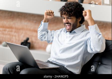 Jeune adulte réussi heureux Indien ou arabe homme d'affaires, courtier ou gestionnaire dans des vêtements formels, assis sur un fauteuil, a eu de bonnes nouvelles, se réjouir de la victoire, bonne affaire,gros profit, gestuelle avec les poings Banque D'Images
