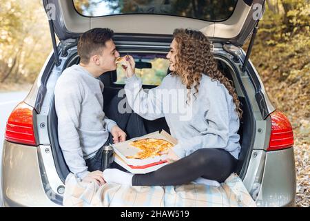 Un homme et une femme mangent de la pizza près de la voiture.Pique-nique à l'automne.Une date romantique pour les amoureux.Nourriture savoureuse. Banque D'Images