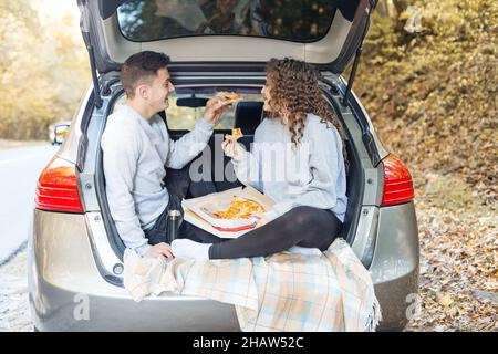 Un homme et une femme mangent de la pizza près de la voiture.Pique-nique à l'automne.Une date romantique pour les amoureux.Nourriture savoureuse. Banque D'Images